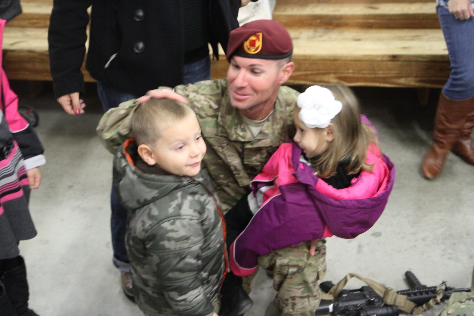 A soldier in a maroon beret kneels to hug his two young children