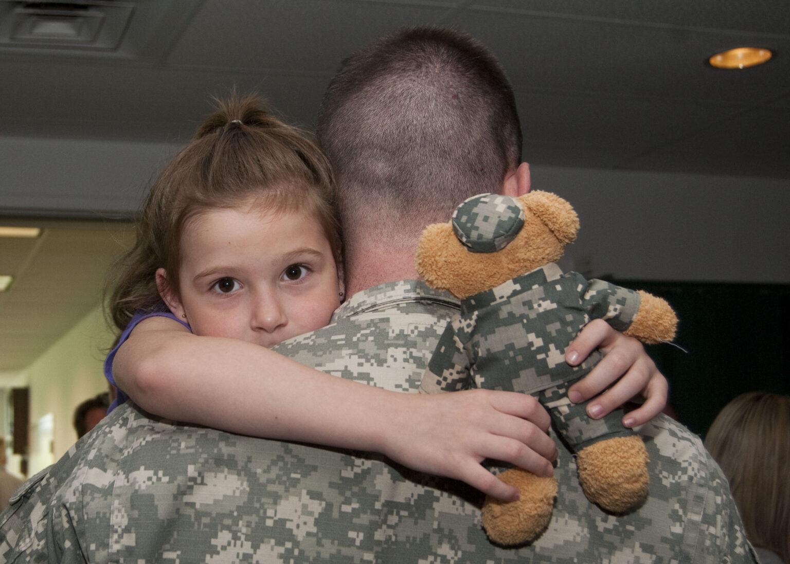A young girl hugs her father, a soldier in uniform, holding a teddy bear dressed in camouflage
